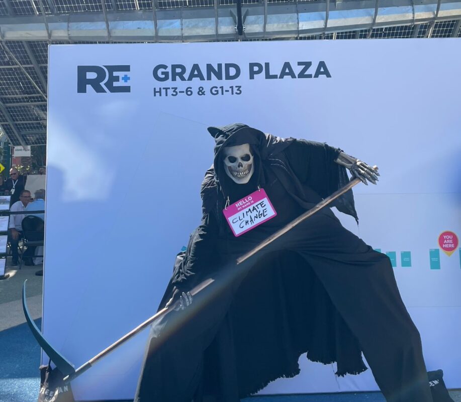 Performer in a Grim Reaper costume stands tall on stilts outside the exhibition hall, with a name tag reading 'Hello, my name is CLIMATE CHANGE'.